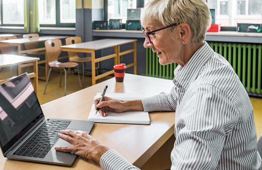 Educator typing on a laptop and writing in a notebook at their desk