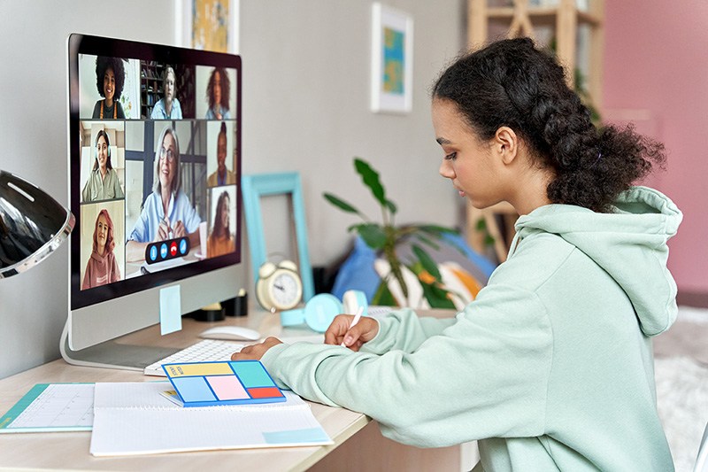 Student writing in a notebook while on a video call with their teacher and class